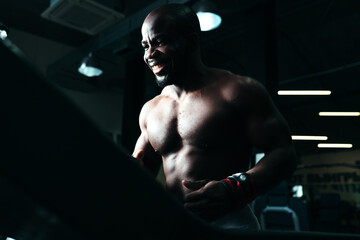 Fototapeta na wymiar An African American man smiles while exercising on a treadmill. Athlete in a naked torso doing a warm-up on the simulator in the gym