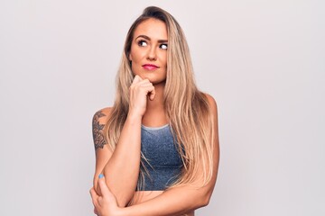 Young beautiful blonde woman wearing casual t-shirt standing over isolated white background thinking concentrated about doubt with finger on chin and looking up wondering