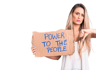 Young beautiful blonde woman holding power to the people cardboard banner with angry face, negative sign showing dislike with thumbs down, rejection concept