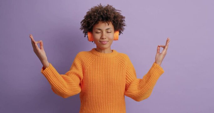 Relaxed calm woman breathes deeply meditates indoor practices yoga after stressful hard day stands with closed eyes wears casual orange jumper wireless headphones models agaist purple background