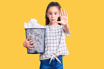 Cute hispanic child girl holding paper bin full of crumpled papers with open hand doing stop sign with serious and confident expression, defense gesture