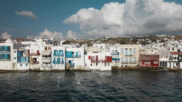 Aerial dolly right shot of white houses on the coast of Mykonos town and a cloudy sky on background
