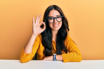 Beautiful brunette young woman wearing glasses and casual clothes sitting on the table smiling positive doing ok sign with hand and fingers. successful expression.