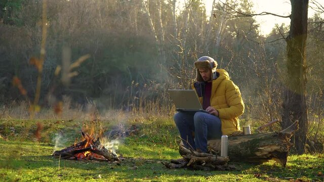 A young man works at a laptop near the fire in the forest. Freelance remote work concept. 4K