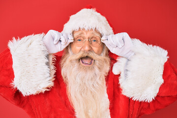 Old senior man with grey hair and long beard wearing santa claus costume holding glasses smiling and laughing hard out loud because funny crazy joke.