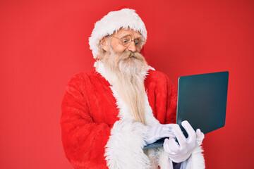 Old senior man with grey hair and long beard wearing santa claus costume using laptop smiling with a happy and cool smile on face. showing teeth.