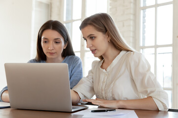 Concentrated female colleagues sit at desk in office look at laptop screen brainstorm over company project. Focused women employees work cooperate using computer. Teamwork, collaboration concept.
