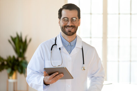 Portrait of smiling young Caucasian male doctor in white medical uniform use tablet in modern hospital or clinic. Happy man GP or physician consult client or patient on pad online. Healthcare concept.