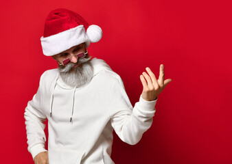 Fototapeta na wymiar Stylish and handsome senior hipster in a Santa hat winks and beckons with his finger calling for New Year's discounts. Man and Santa hat stands on a red background and looks away. Place for text.