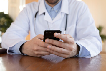 Crop close up of male doctor in white medical uniform sit at desk in clinic use modern cellphone gadget. Man GP or physician hold smartphone consult patient or client online in modern hospital.