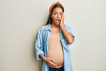 Beautiful hispanic woman expecting a baby, touching pregnant belly afraid and shocked, surprise and...