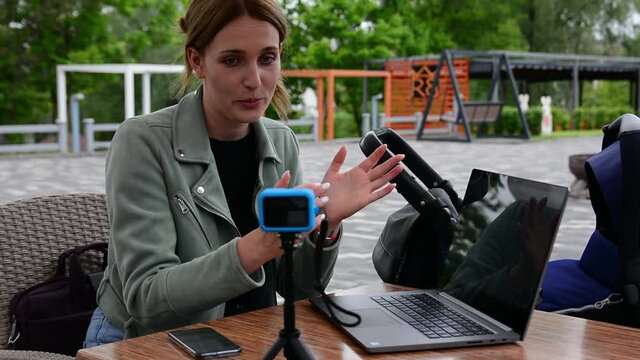 Girl blogger blogging in a cafe on a summer terrace outdoors. A blogger mom with a baby in a pram shoots her vlogs and short videos on an action camera on a tripod, and stylish laptop on a table