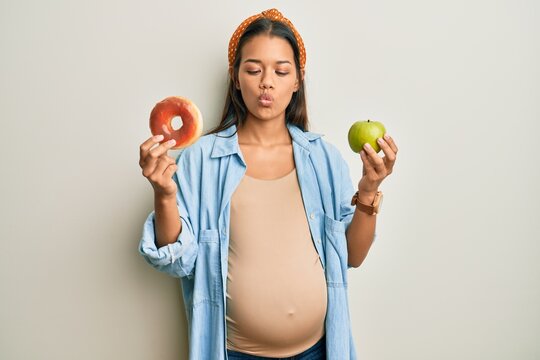 Beautiful hispanic woman expecting a baby, holding food making fish face with mouth and squinting eyes, crazy and comical.