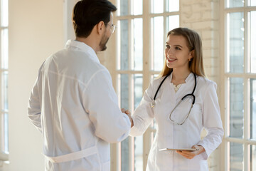 Smiling diverse doctors in white medical uniforms shake hands greeting get acquainted in hospital. Happy male and female physicians handshake close agreement. Healthcare, recruitment concept.