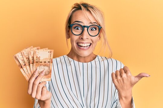 Beautiful blonde woman holding south african 20 rand banknotes pointing thumb up to the side smiling happy with open mouth