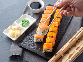 Philadelphia and California rolls on a textured dark plate. One roll is held in Granny sticks above the others. Next to soy sauce, wasabi and ginger. Side view. Japanese cuisine