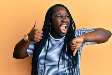 Young black woman with braids doing thumbs down and thumbs up gesture angry and mad screaming...