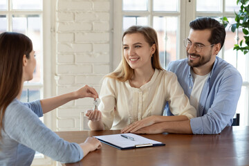 Female real estate agent give keys to excited young Caucasian couple renters buying first home...