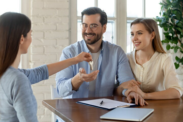 Female relator or broker give keys to new house to excited Caucasian couple renters at office meeting. Woman real estate agent congratulate spouses with home purchase. Ownership concept.