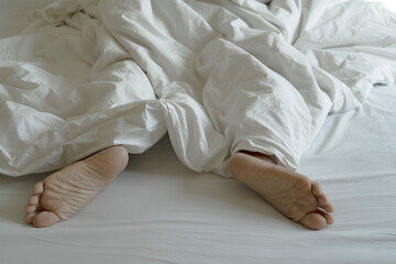 The feet or foot of couples sleeping on the bed and cover by the white blanket on a bright Sunday morning