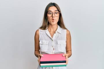 Young hispanic woman holding books making fish face with mouth and squinting eyes, crazy and comical.
