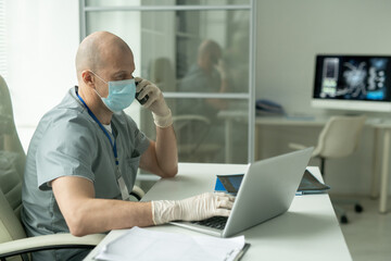 Side view of mature male clinician in mask sitting by desk in front of laptop