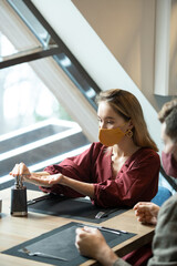 Young female in protective mask taking sanitizer t owash hands before dinner