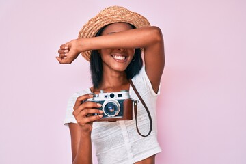 Young african american woman wearing summer hat holding vintage camera smiling cheerful playing peek a boo with hands showing face. surprised and exited