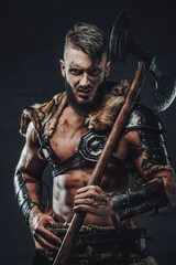 Fototapeta premium Northern savage warrior with muscular build staring at camera holds his two handed axe on his shoulder in dark background.