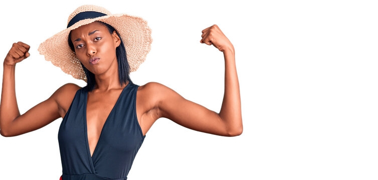 Young african american woman wearing swimsuit and summer hat showing arms muscles smiling proud. fitness concept.