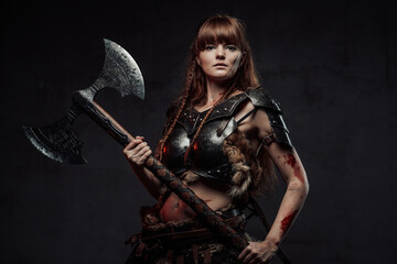 Fototapeta na wymiar Wielding two handed axe grimy nordic amazon with brown hairs in dark armour posing in dark background.