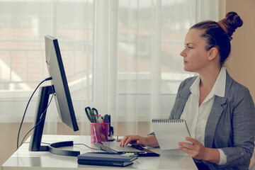 Side view profile of thoughtful young woman sitting near laptop and holding a notebook.