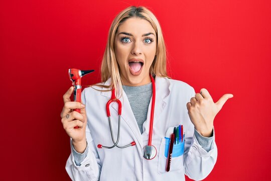 Young caucasian woman wearing doctor uniform holding otoscope pointing thumb up to the side smiling happy with open mouth
