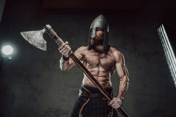 Muscular and furious nord warrior staying with naked torso with helmet on his head holding his two...