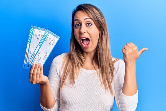 Young beautiful woman holding airline boarding pass pointing thumb up to the side smiling happy with open mouth