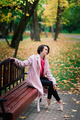 Young beautiful ballerina in fair coat and pointe shoes sit on the bench, rest outdoors.