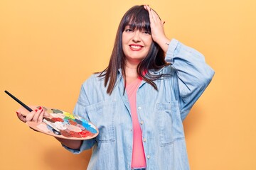 Young plus size woman holding paintbrush and palette stressed and frustrated with hand on head,...