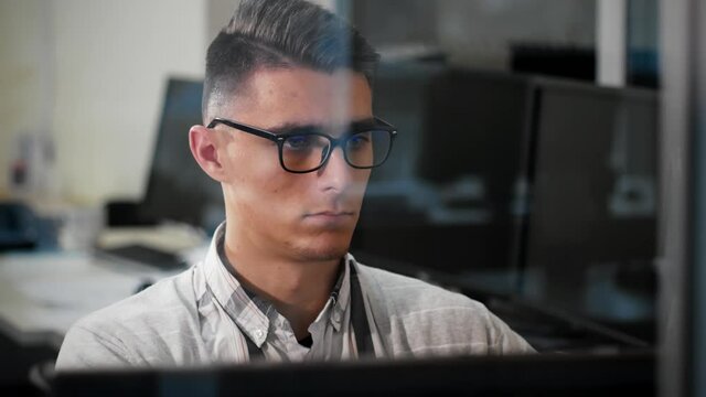 Portrait video shooting through the window of a young man in glasses working at a computer at his workplace in the office. Reflections of passers-by and passing cars are visible in the window glass.