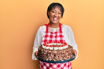 Middle age african american woman wearing baker apron holding homemade cake winking looking at the...