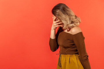 Side view portrait of unhappy young woman with blonde curly hair in casual clothes standing and covering with hand on head, feeling sorrow. Indoor studio shot isolated on red background, copy space