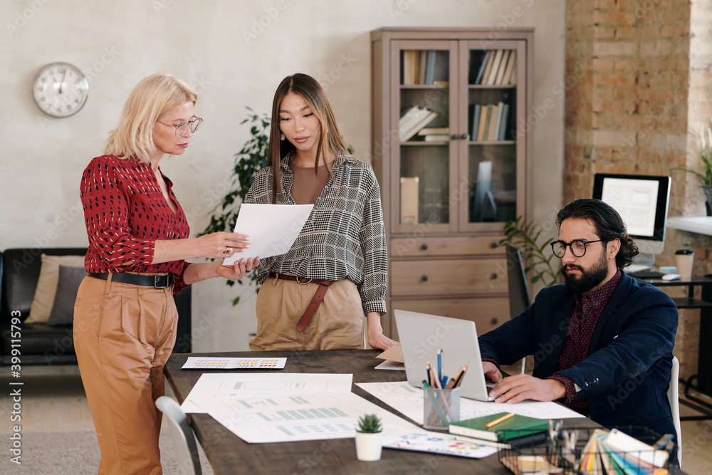 Poster young and mature females standing by table and discussing sketch on paper