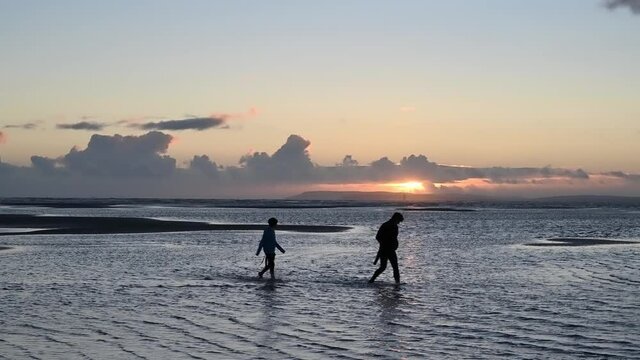 Two people silhouetted by a beautiful sunset while walking in the sea at low tide at West Wittering in West Sussex.