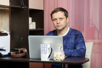 A man in home clothes works at a laptop. Looks into the camera.