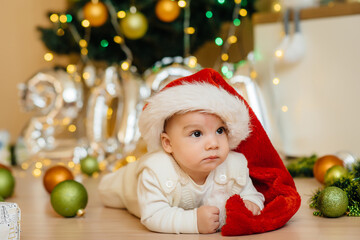 Cute smiling baby is lying under a festive Christmas tree and playing with gifts. Christmas and New Year celebrations