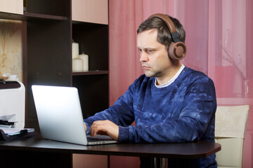 A man in home clothes works at a laptop. Headphones are on the head.