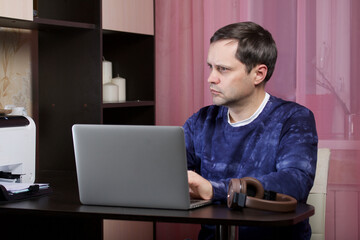 A man in home clothes works at a laptop. Headphones are on the table nearby.
