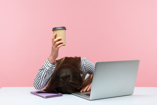 Sleepy bored woman office worker lying on table with laptop holding and showing paper coffee cup, feeling lack of energy sitting at workplace. Indoor studio shot isolated on pink background