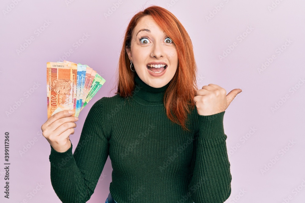 Sticker beautiful redhead woman holding south african rand banknotes pointing thumb up to the side smiling h