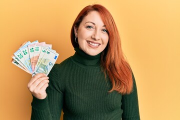 Beautiful redhead woman holding euro banknotes looking positive and happy standing and smiling with a confident smile showing teeth