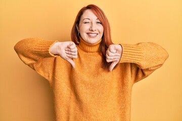 Beautiful redhead woman doing negative thumbs down gesture winking looking at the camera with sexy...